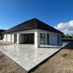 Nowoczesny dom jednorodzinny z rozległym tarasem wyłożonym jasnoszarą, błyszczącą płytką ceramiczną, widok z zewnątrz w słoneczny dzień.