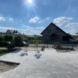 Nowo ułożona, szara kostka brukowa na podjeździe do domu jednorodzinnego, z przygotowanym miejscem na rabatę, widok na ogród i ogrodzenie.