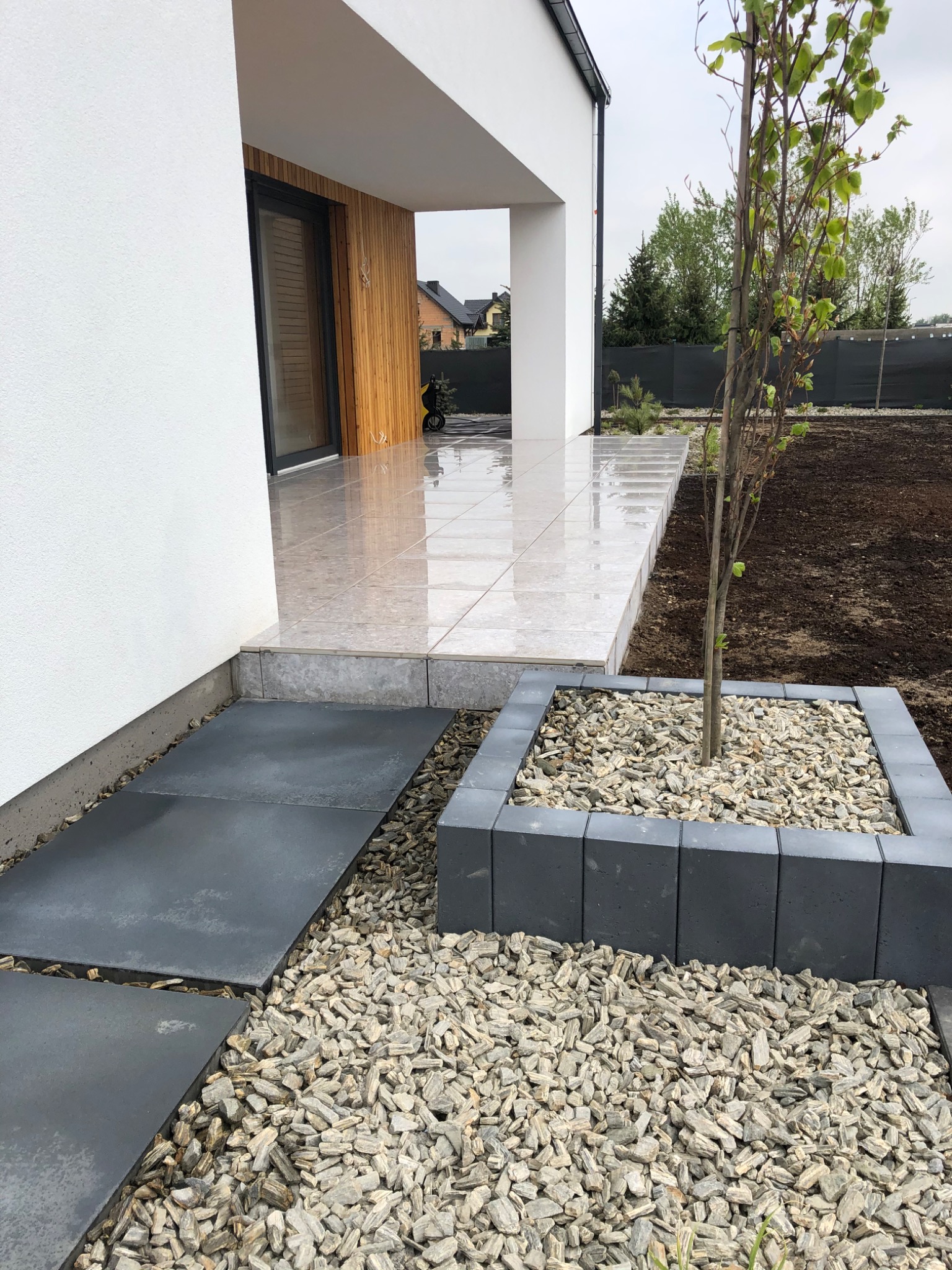 Nowoczesny taras z płytek ceramicznych, ciemnoszare płyty chodnikowe oddzielone korą, minimalistyczna donica z młodym drzewkiem na tle białej elewacji domu z elementami drewnianymi.