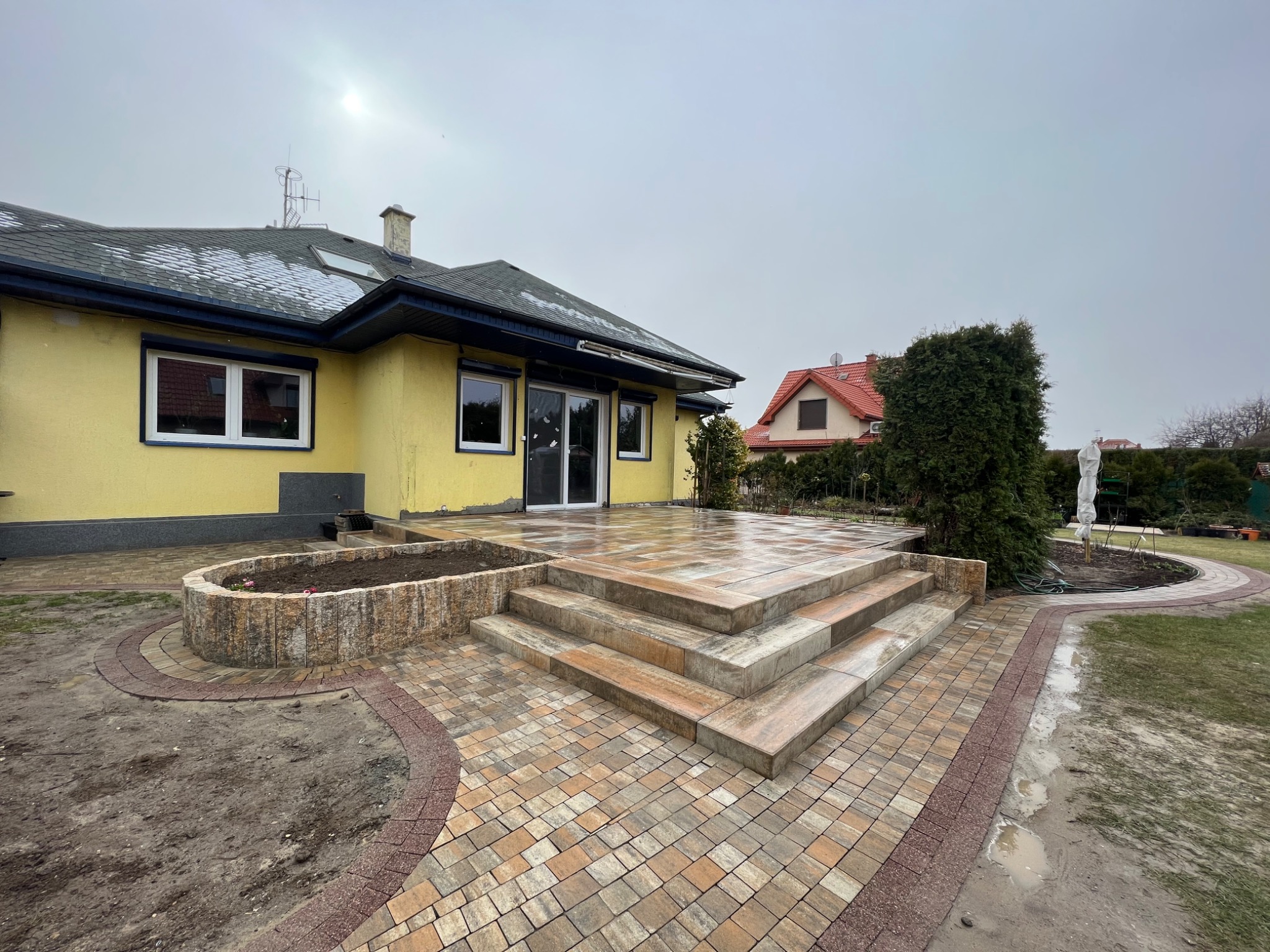 Wykończony taras z kostki brukowej w odcieniach beżu i brązu, z trzema schodami prowadzącymi do domu z żółtą elewacją i szarym dachem, widok na ogród i sąsiedni dom w tle.