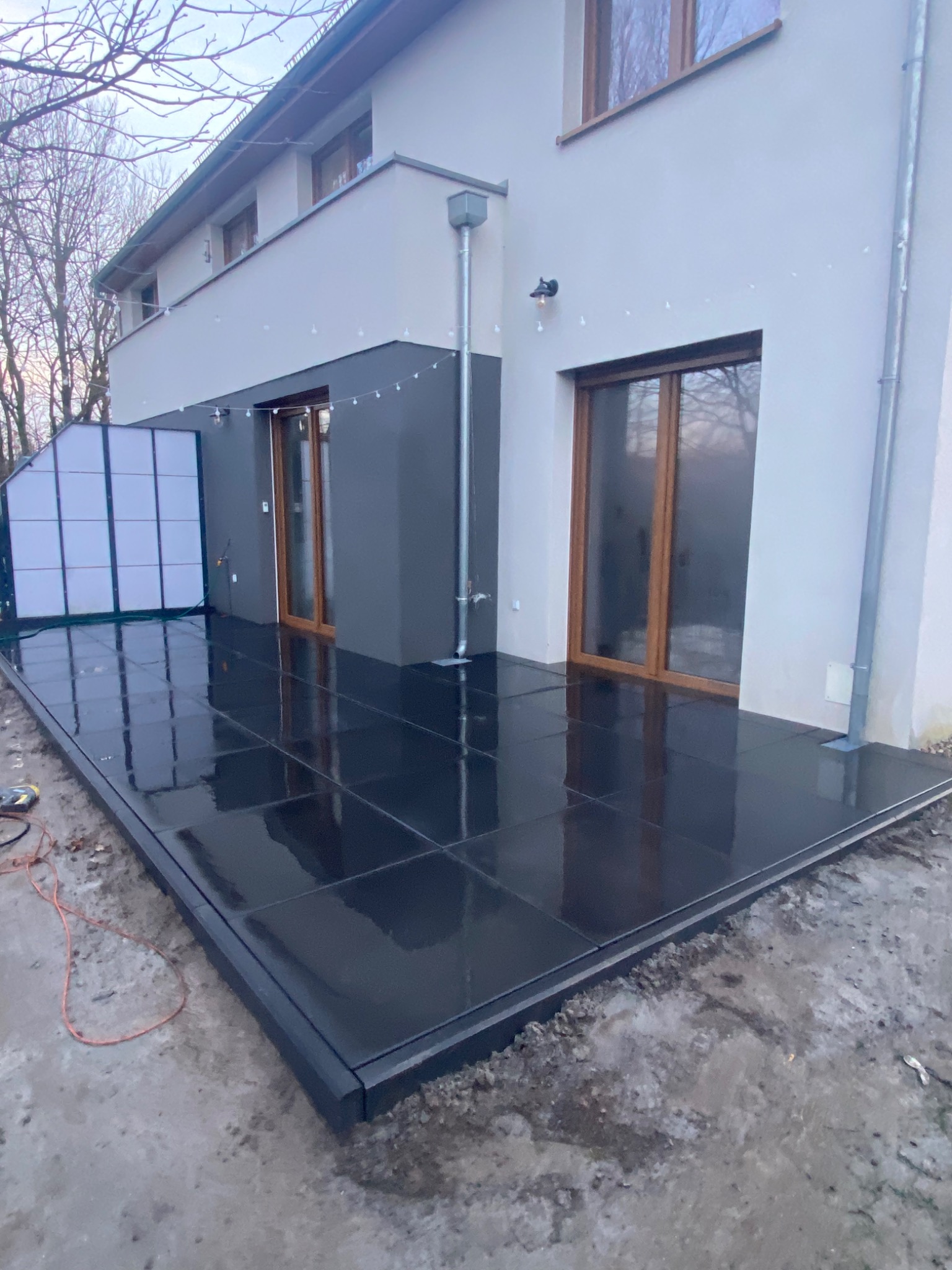 Czarny taras z płytek ceramicznych przy nowoczesnym domu, z widoczną konstrukcją zadaszenia po lewej stronie i elementami wykończeniowymi w trakcie prac.