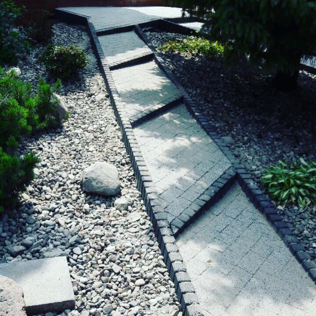 Ścieżka z kostki brukowej prowadząca przez ogród, otoczona drobnymi kamieniami i zielenią, z widocznym spadkiem terenu.