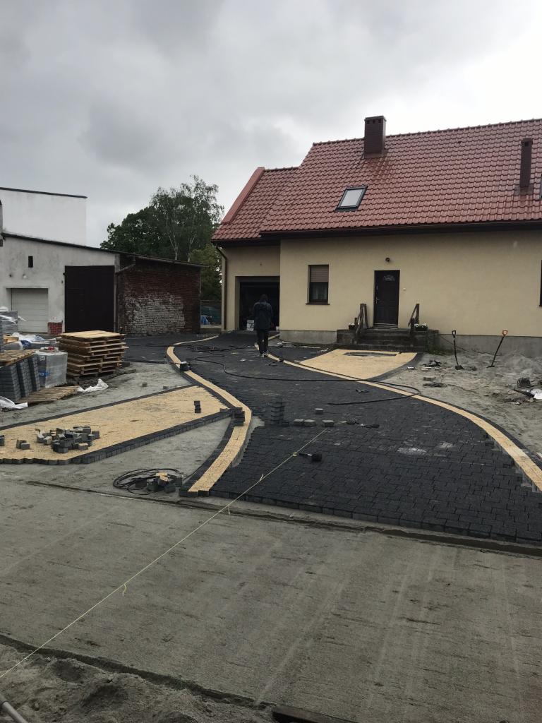 Częściowo ułożona, kręta ścieżka z ciemnej kostki brukowej z jasną obwódką prowadząca do domu jednorodzinnego; widoczne materiały budowlane i narzędzia pracy.
