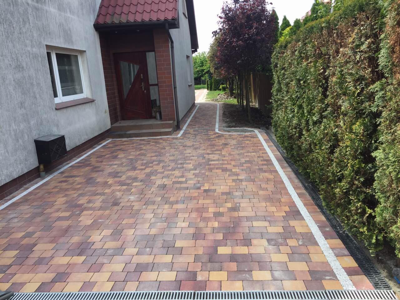 Kolorowa kostka brukowa ułożona na podjeździe przed domem, z obrzeżem z jasnego żwiru i systemem odprowadzania wody.