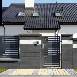 Nowoczesne ogrodzenie panelowe z bramą wjazdową, wykonane z szarych prefabrykatów betonowych i poziomych paneli, z widocznym domem z ciemnym dachem w tle.