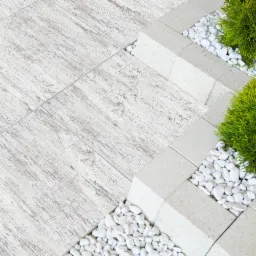 Jasnoszara nawierzchnia z płytek imitujących drewno, obrzeże z jasnych, betonowych kostek, wypełnienie białym grysem i zielonymi krzewami.