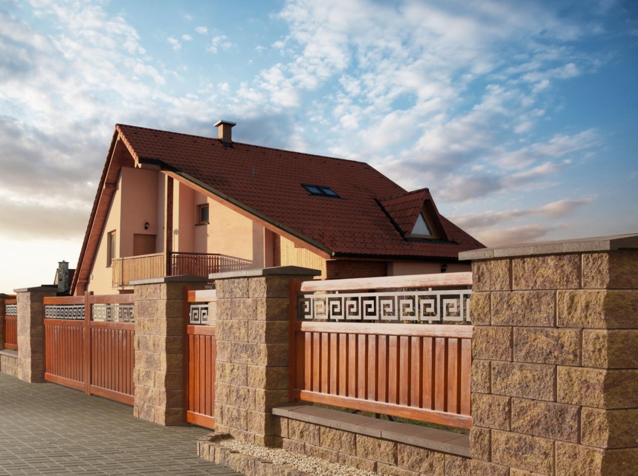 Nowoczesne ogrodzenie z drewnianymi przęsłami i kamiennymi słupkami w Jaczowie, z dekoracyjnym wzorem meandrycznym, na tle domu z czerwoną dachówką i błękitnego nieba.