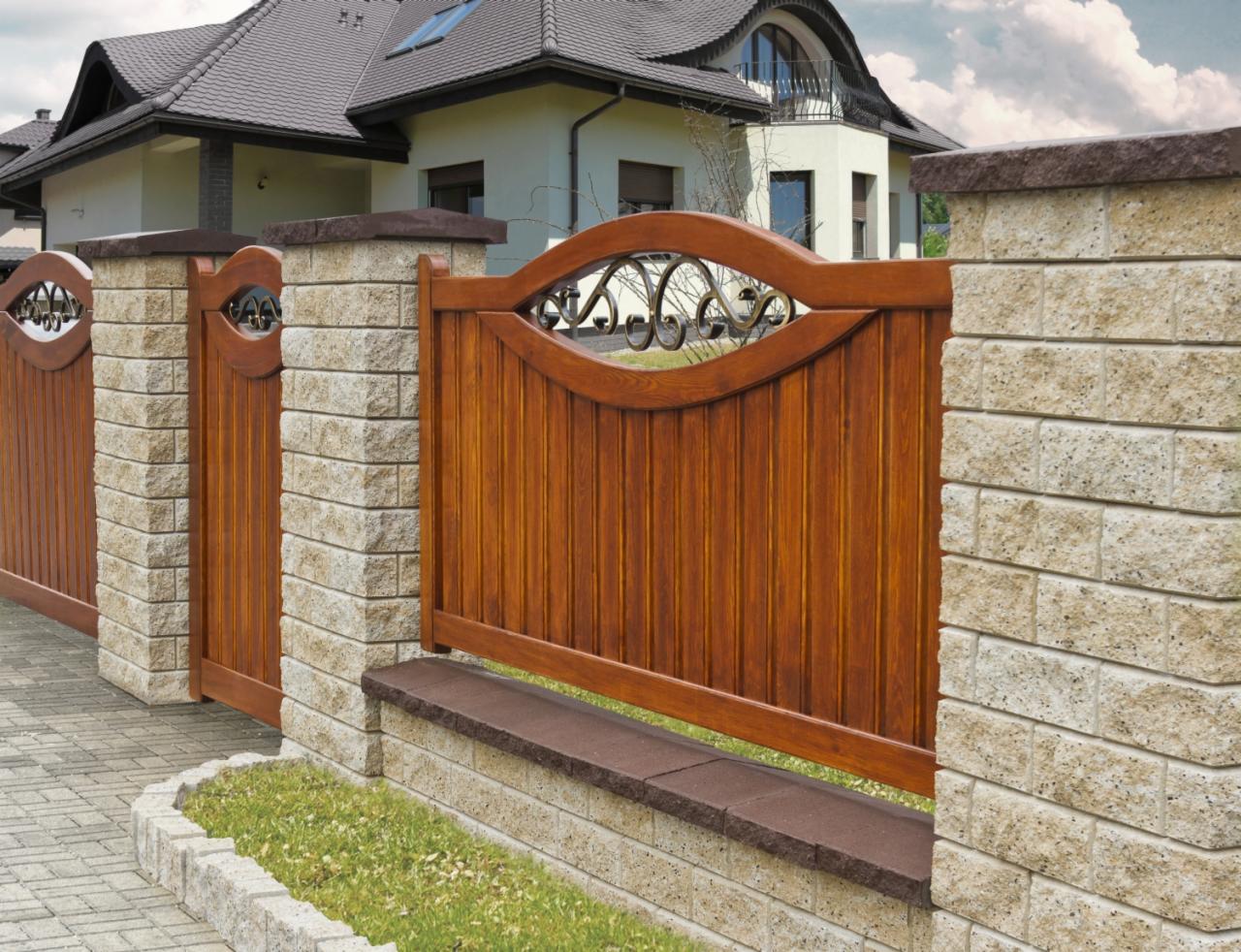 Ozdobne drewniane przęsło ogrodzenia z metalowymi elementami dekoracyjnymi, umieszczone pomiędzy słupkami z jasnego kamienia łupanego, widok z poziomu chodnika.