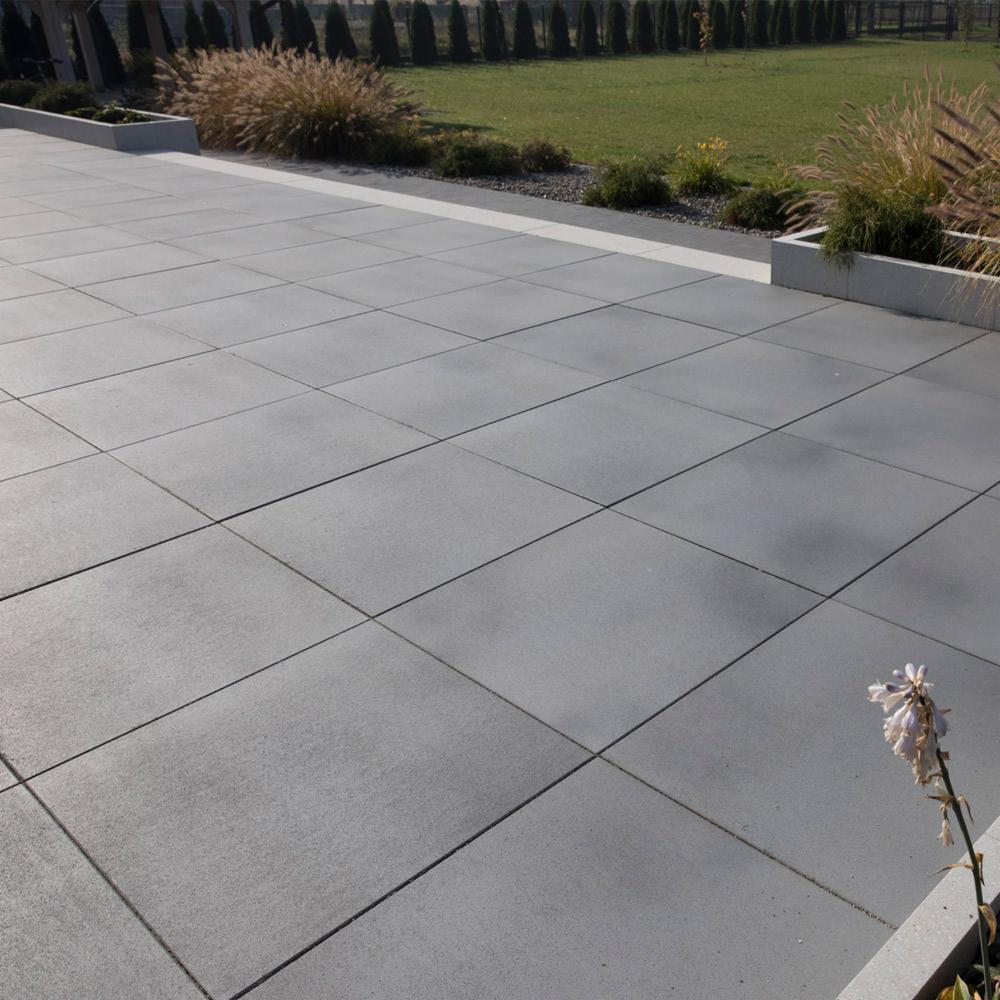 Szara nawierzchnia z dużych betonowych płyt ułożonych w kwadraty, z widocznymi fugami, otoczona zielonym trawnikiem i ozdobnymi trawami w betonowych donicach, z pojedynczym przekwitłym kwiatem w rogu.