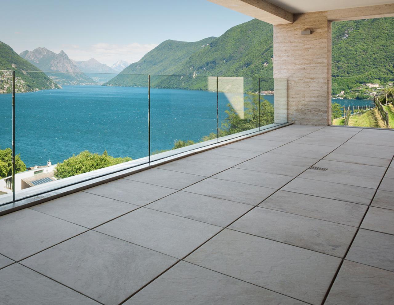 Przestronny taras z szarych płyt kamiennych, zabezpieczony szklaną balustradą z widokiem na malownicze jezioro i góry w oddali.