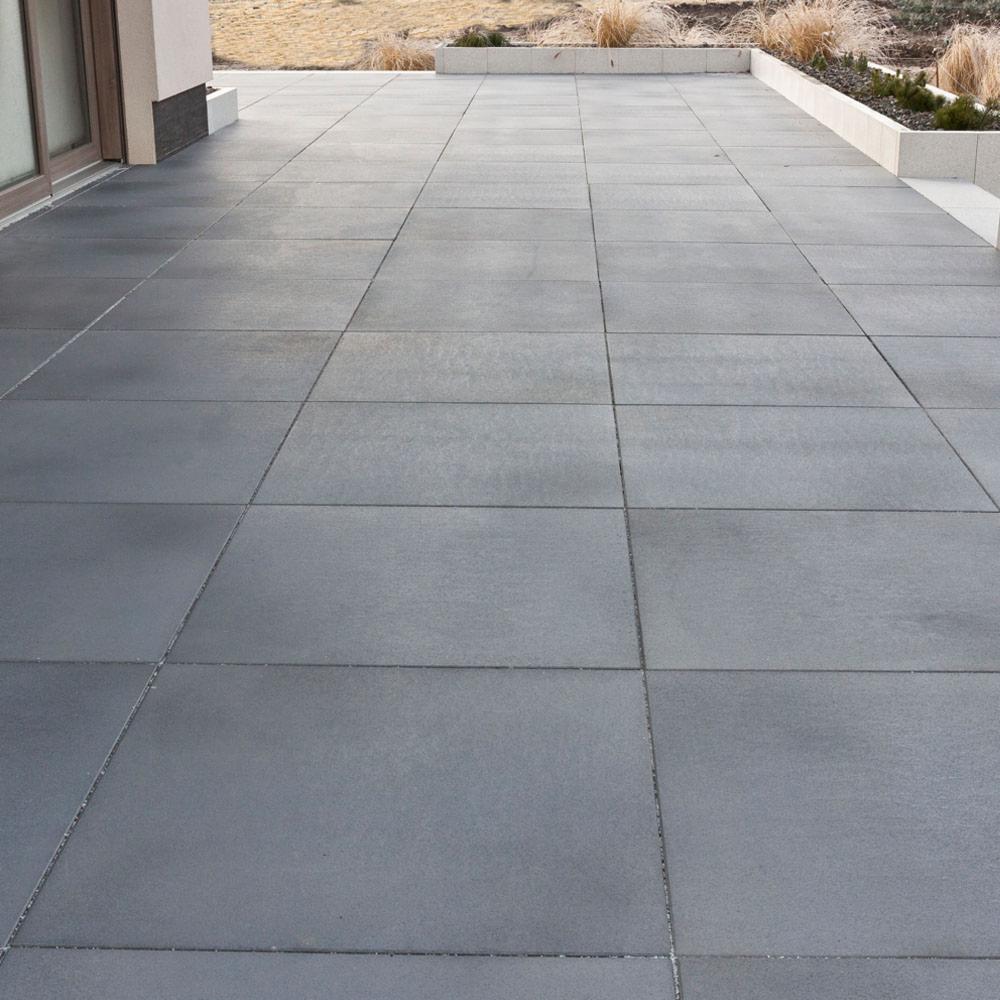 Nowoczesny taras z dużych, szarych płyt betonowych, z widocznym przeszkleniem drzwi balkonowych i rabatą z suchymi trawami w tle.