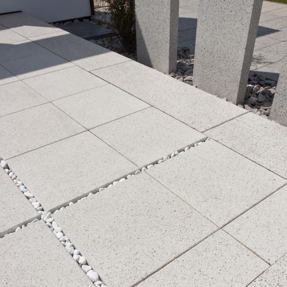 Jasnoszara nawierzchnia z kwadratowych płyt betonowych z białymi kamykami w fugach, prowadząca do dwóch słupków z tego samego materiału, z roślinnością w tle.