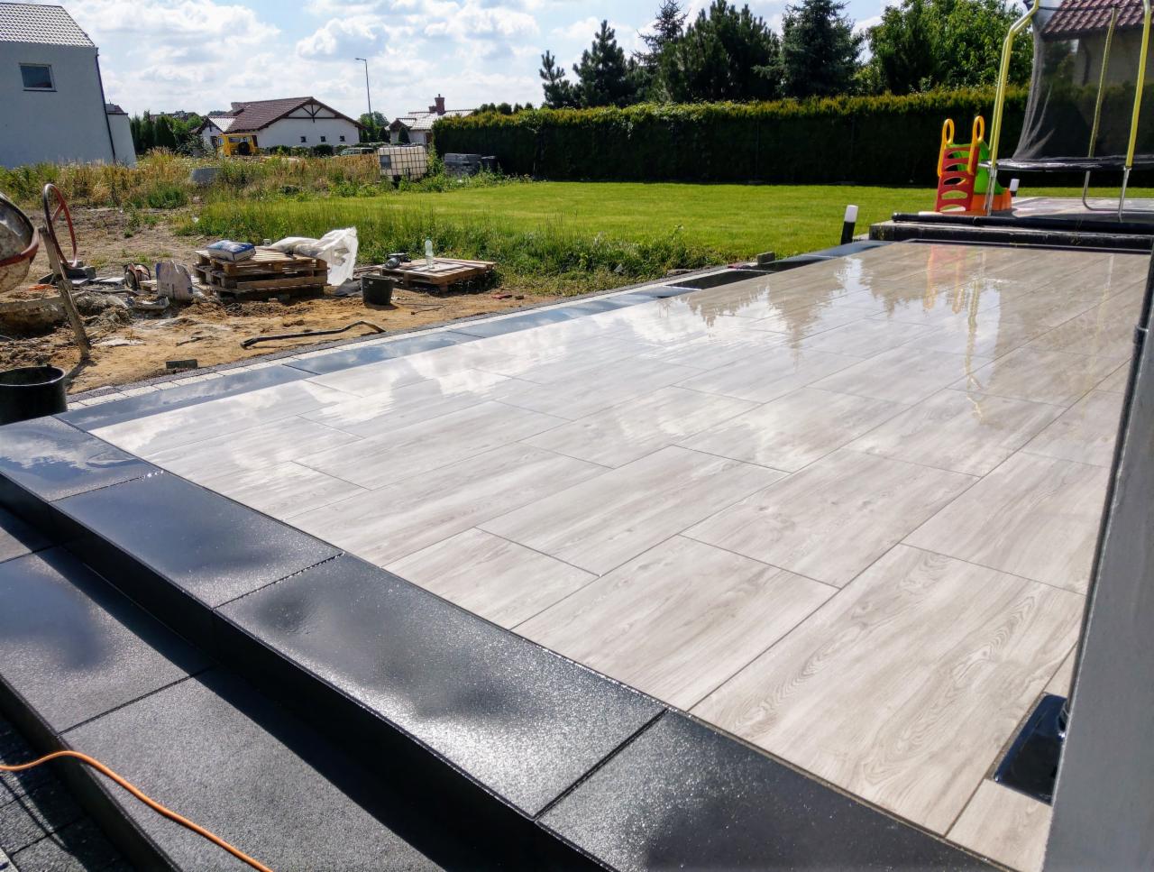 Świeżo ułożony taras z jasnych płyt imitujących drewno, obramowany ciemnoszarą kostką, widoczne odbicia chmur na mokrej powierzchni, w tle trawnik, trampolina i domy.