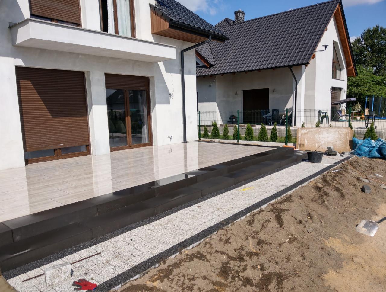 Nowoczesny taras z jasnych płytek ceramicznych i czarnych stopni prowadzących do domu, obok ułożona nawierzchnia z jasnoszarej kostki brukowej z czarnym obramowaniem, w tle dom z ciemnym dachem...