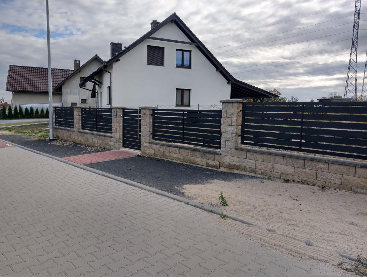 Nowoczesne, poziome, czarne ogrodzenie z paneli zamontowane na niskim murku z jasnego kamienia, z bramą wjazdową, na tle białego domu z brązowym dachem i szarym niebem.