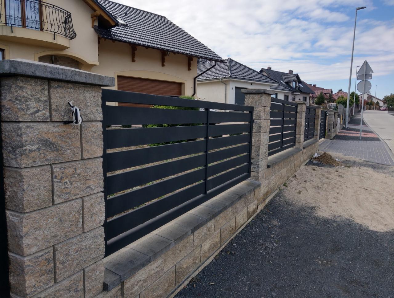 Nowoczesne ogrodzenie z poziomych, czarnych paneli zamocowanych między słupkami z kamiennej kostki, widok z zewnątrz posesji, z fragmentem chodnika i ulicy w tle.