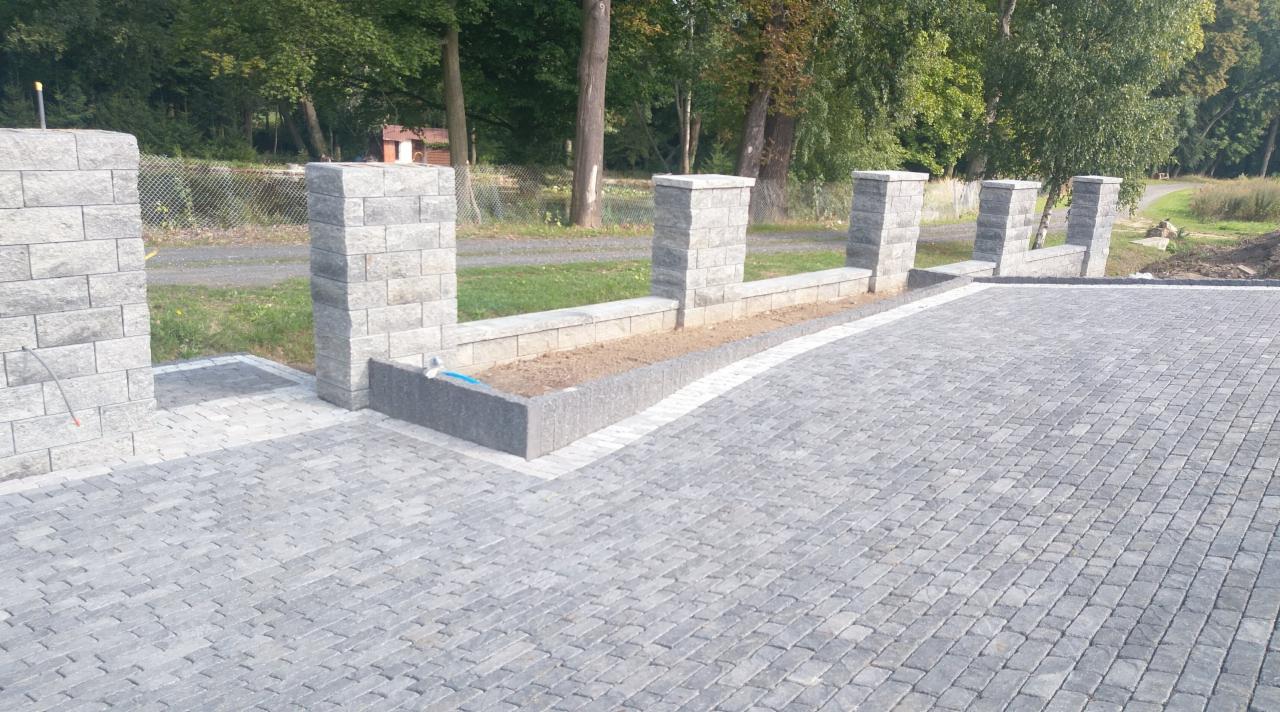 Ułożona szara kostka betonowa tworząca podjazd z niskim murem oporowym z prefabrykatów betonowych oraz słupkami z tego samego materiału, w tle zieleń drzew.