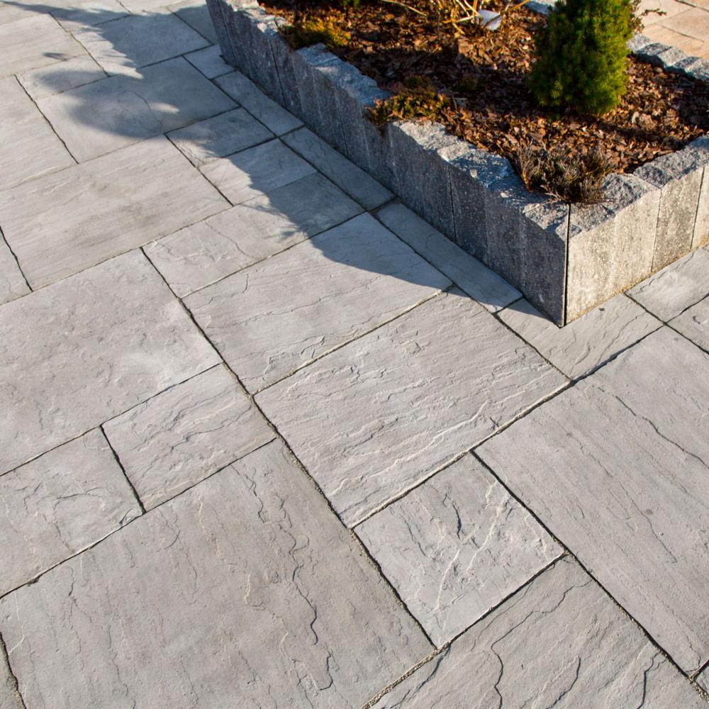 Szara nawierzchnia z nieregularnych płyt kamiennych ułożonych wokół podwyższonej rabaty z kamienną obudową, wypełnionej korą i niskimi roślinami iglastymi.