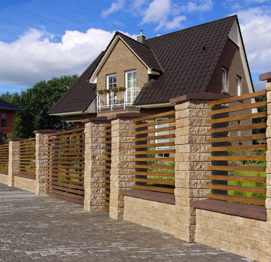 Nowoczesne ogrodzenie z kamiennych słupków i drewnianych, poziomych przęseł na tle domu z brązową dachówką i białym balkonem.