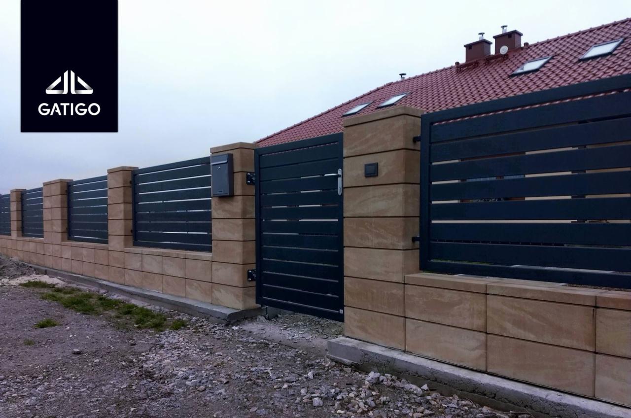 Nowoczesne ogrodzenie z poziomych paneli w kolorze antracytu, osadzone na murku z beżowych bloczków, z przesuwną bramą wjazdową i skrzynką na listy, na tle domu z czerwoną dachówką