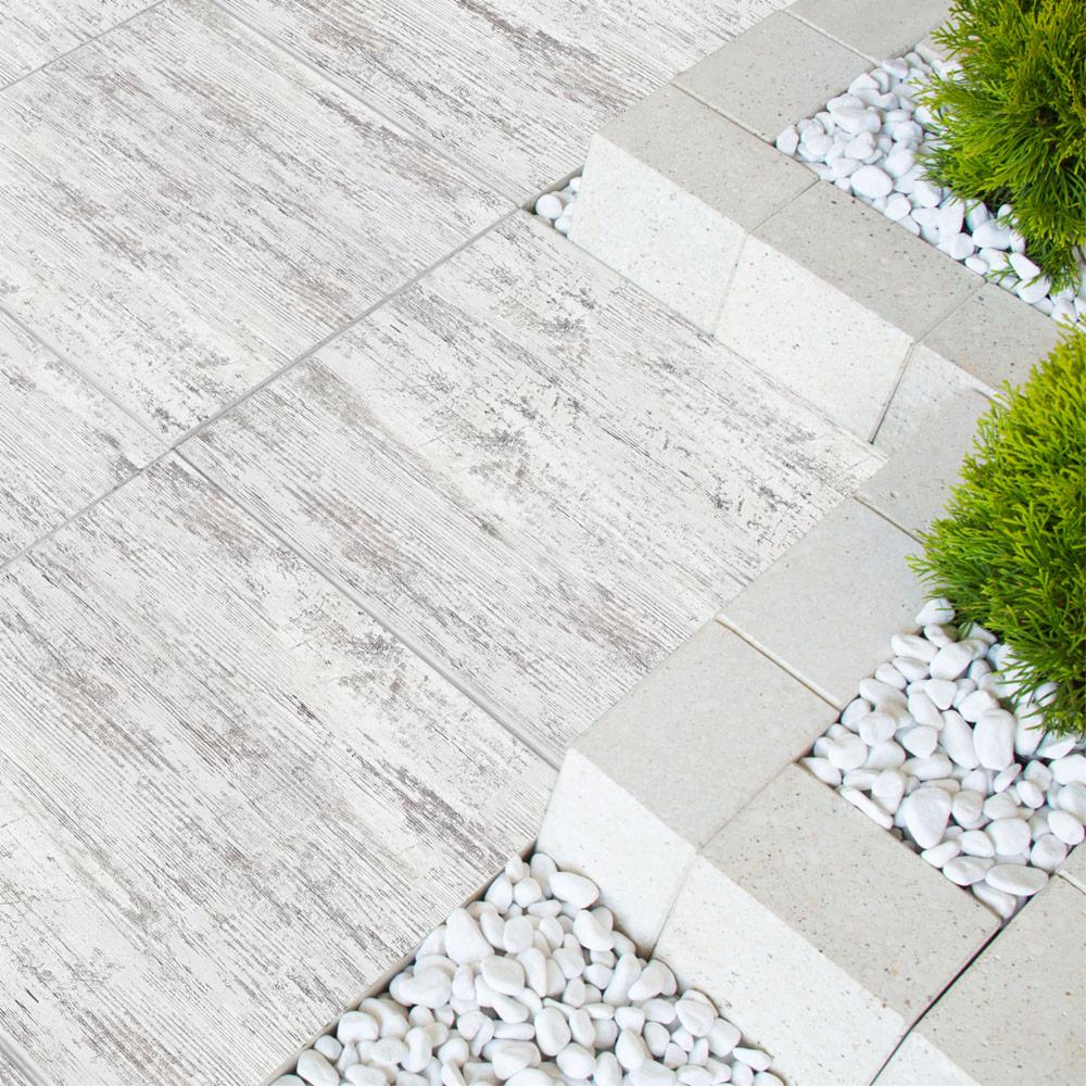 Jasnoszara nawierzchnia z płytek imitujących drewno, obrzeże z jasnych, betonowych kostek, wypełnienie białym grysem i zielonymi krzewami.