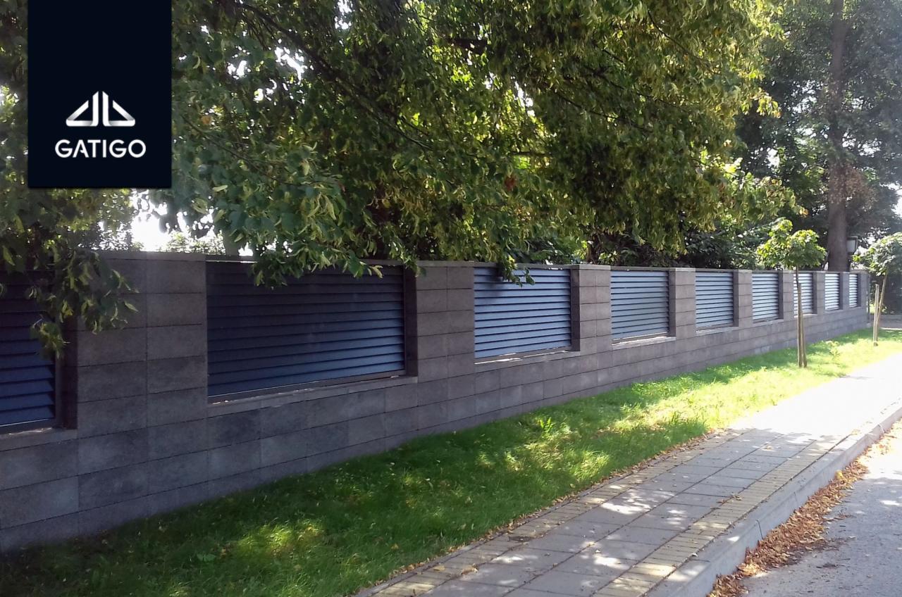 Długi, nowoczesny płot z szarych betonowych bloków i poziomych, ciemnoszarych paneli, widoczny z perspektywy chodnika, z zielenią w tle.
