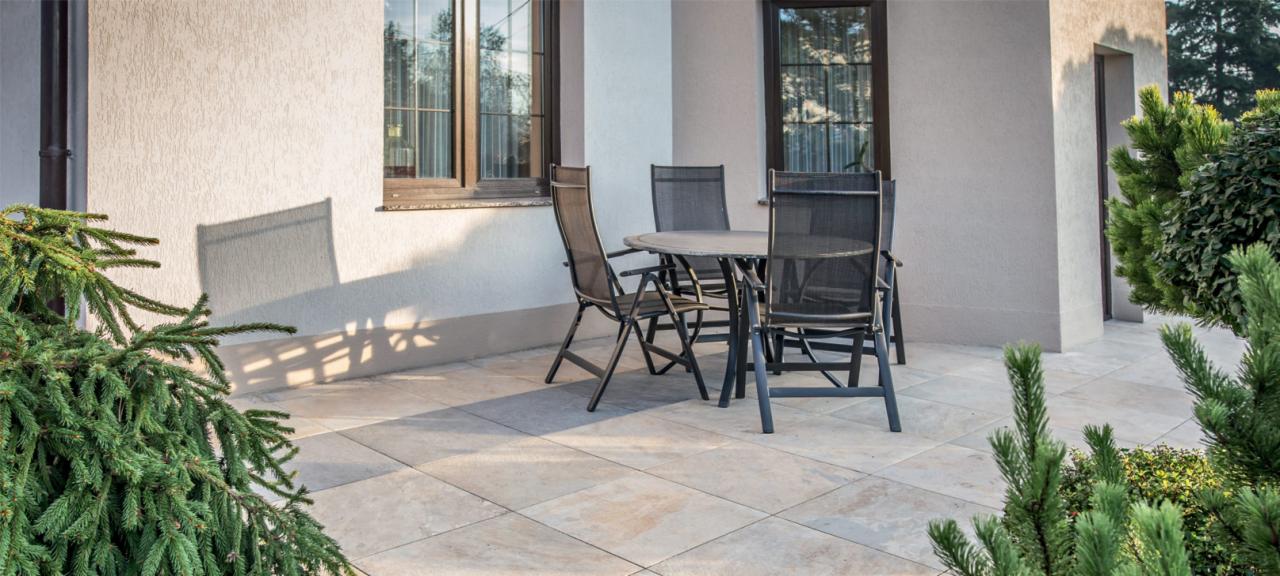 Jasny taras wyłożony kwadratowymi płytami, zestaw mebli ogrodowych z czarnymi krzesłami i okrągłym stołem, ściana budynku z oknem i fragmentem iglaka.