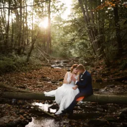 Para młoda siedząca na powalonym drzewie nad strumieniem w lesie, panna młoda w białej sukni z koronkową górą i błyszczącymi butami, pan młody w granatowym garniturze, jesienne liście na ziemi...