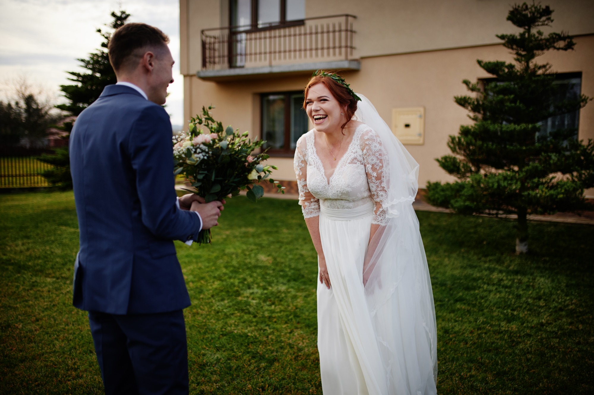 Uśmiechnięta panna młoda w koronkowej sukni i wianku na głowie, z bukietem wręczanym przez pana młodego w granatowym garniturze, na tle domu i trawnika.