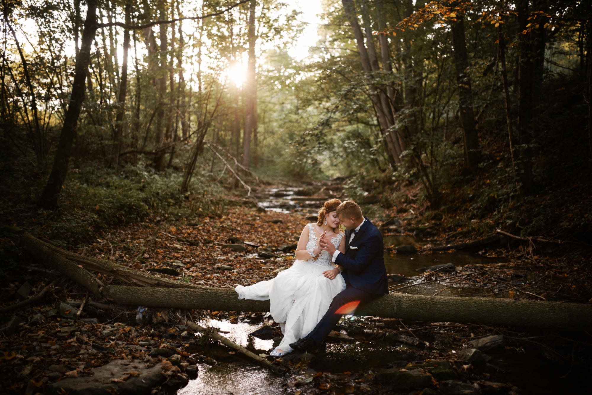 Para młoda siedząca na powalonym drzewie nad strumieniem w lesie, panna młoda w białej sukni z koronkową górą i błyszczącymi butami, pan młody w granatowym garniturze, jesienne liście na ziemi...
