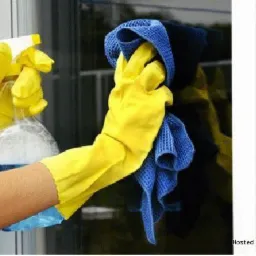 Czyszczenie okna z zewnątrz: ręce w żółtych rękawicach spryskują szybę płynem i wycierają niebieską ściereczką z mikrofibry, widoczna biała elewacja budynku.