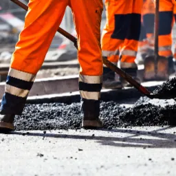 Drogowcy w pomarańczowych kombinezonach wyrównują asfalt na drodze, jeden z nich z łopatą w dłoni