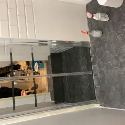 Nowoczesna łazienka z ciemną heksagonalną podłogą, białymi prostokątnymi płytkami na ścianie i szklaną kabiną prysznicową, widoczne butelki ze środkami czystości.