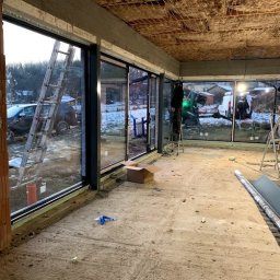 MAST - Śląskie Okna - Montaż przesuwnych okien w surowym wnętrzu. Widoczne drabiny, narzędzia i pracownik na drabinie. Na zewnątrz widok na zimowy krajobraz.