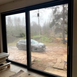 MAST - Śląskie Okna - Duże przesuwne okno z czarną ramą, widok na zewnątrz z zaparkowanym samochodem. Wnętrze w stanie surowym, widoczne płyty i kartony na podłodze.