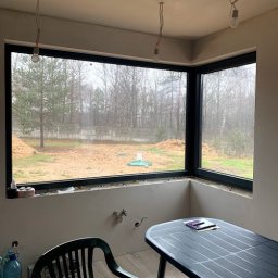 MAST - Śląskie Okna - Narożne okno z czarną ramą w surowym wnętrzu, widok na ogród z drzewami. Stolik i krzesło w pomieszczeniu. Niedokończone wykończenie ścian.