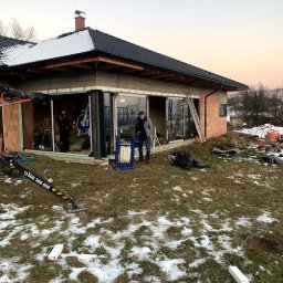 MAST - Śląskie Okna - Montaż przesuwnych szklanych drzwi tarasowych w surowym stanie budynku. Widoczny pracownik z podnośnikiem, drabina i ekipa montażowa wewnątrz.