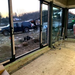 MAST - Śląskie Okna - Montaż przesuwnych okien z czarnymi ramami. Na zewnątrz widoczny samochód z dźwigiem podczas instalacji. Wnętrze w stanie surowym.