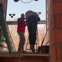 MAST - Śląskie Okna - Montaż dużego okna przez dwóch monterów za pomocą przyssawki próżniowej w surowym stanie budynku z cegły ceramicznej. Widoczne drabiny i narzędzia.