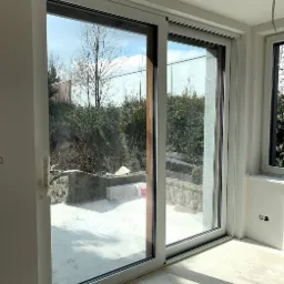 Nowoczesne, przesuwne okno tarasowe z białymi ramami, widok na ogród. Elementy wykończeniowe w kolorze drewna. Jasne wnętrze w trakcie prac wykończeniowych.