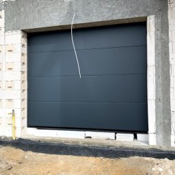 MAST - Śląskie Okna - Szara segmentowa brama garażowa w surowym otworze budynku z białego bloczka, widoczny przewód instalacyjny i żółty zawór gazowy.