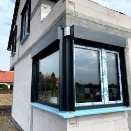 MAST - Śląskie Okna - Narożne okno z roletami zewnętrznymi w surowym budynku, widoczne nieotynkowane ściany z betonu komórkowego i fragmenty innych okien na wyższych kondygnacjach.