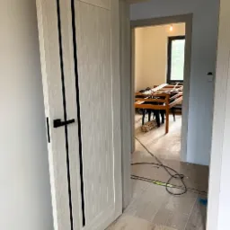 Otwarta, jasna dębowa drzwi wewnętrzne z czarnymi, pionowymi wstawkami i klamką, widok na wnętrze z drewnianym stołem i oknem. Podłoga wyłożona płytkami imitującymi drewno.