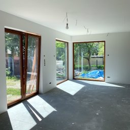 MAST - Śląskie Okna - Wnętrze w stanie deweloperskim z dużymi oknami drewnianymi i przesuwnymi drzwiami balkonowymi. Widoczne przygotowania do wykończenia, surowy beton na podłodze.