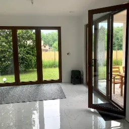 Nowoczesne, brązowe drzwi balkonowe otwarte na taras. Widok na ogród przez duże okno. Białe marmurowe płytki podłogowe. W tle zielony taboret.