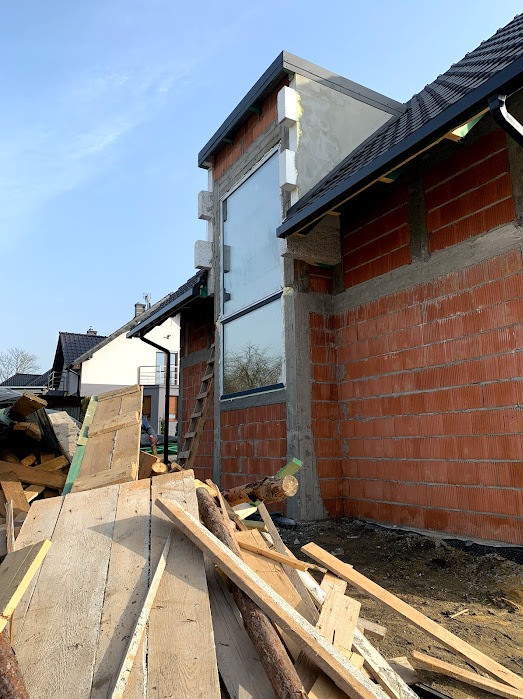 Montaż okna w surowym budynku z czerwonej cegły. Widoczna drabina i stos drewna. Okno duże, prostokątne z czarną ramą. Budynek w trakcie budowy, częściowo otynkowany.