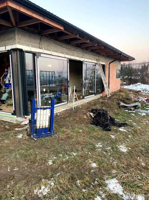 Montaż dużych okien przesuwnych w surowym budynku. Widoczny sprzęt montażowy i drabina. Zewnętrzna strona budynku w trakcie prac wykończeniowych.