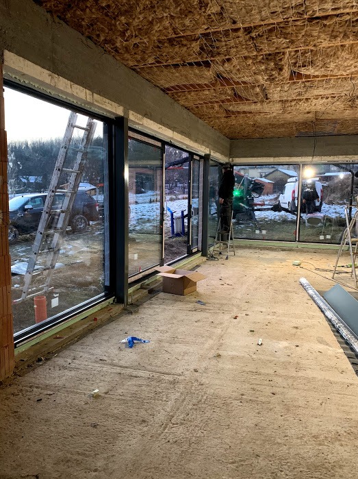 Montaż przesuwnych okien w surowym wnętrzu. Widoczne drabiny, narzędzia i pracownik na drabinie. Na zewnątrz widok na zimowy krajobraz.