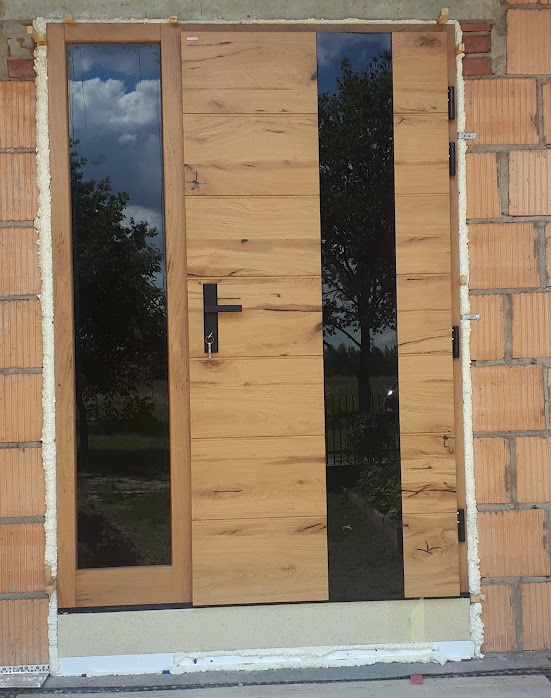 Nowoczesne drzwi zewnętrzne z drewna i szkła w surowym murze. Widoczne detale wykończenia i montażu, odbicie drzewa w szklanych panelach.