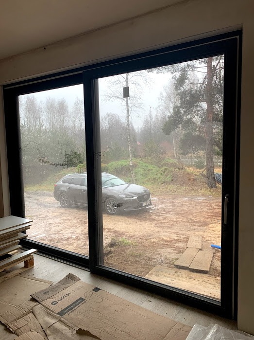 Duże przesuwne okno z czarną ramą, widok na zewnątrz z zaparkowanym samochodem. Wnętrze w stanie surowym, widoczne płyty i kartony na podłodze.